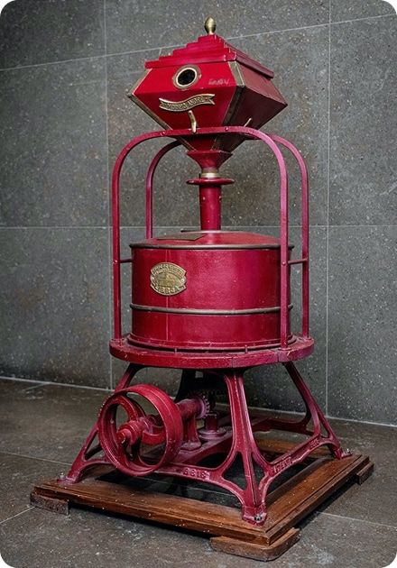 A tall, ornate dark red vintage machine with a faceted hopper top, cylindrical midsection, and a wheeled cast iron base.