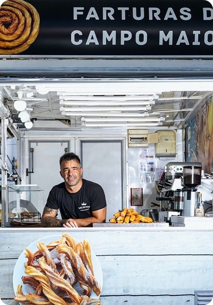 A man smiles from inside a food stall displaying and serving "farturas" (Portuguese fried dough pastries), with a large plate of them in the foreground.