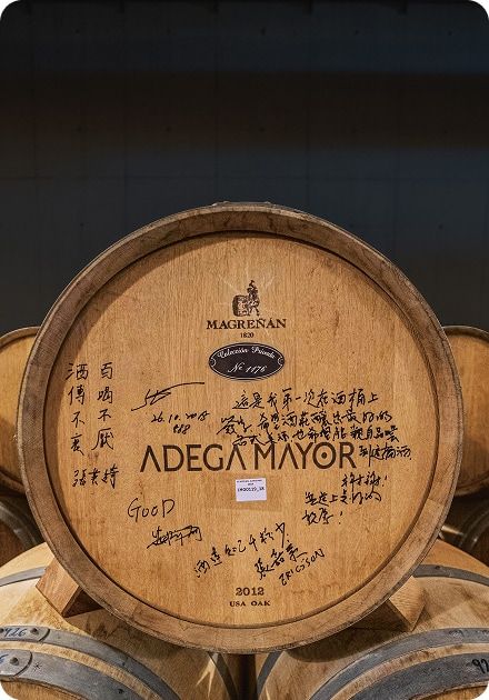 A wooden wine barrel with Magreñan and Adega Mayor branding, covered in handwritten signatures and messages, some in Chinese.