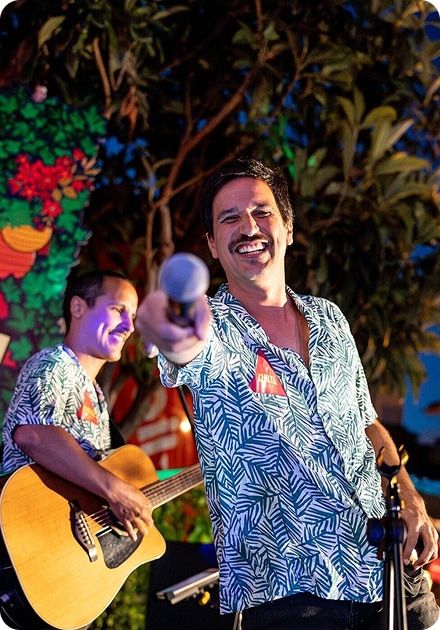 Two smiling men in matching tropical shirts perform outdoors at night, one holding a microphone towards the viewer and the other playing guitar.