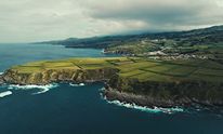 Aerial view of lush green coastal cliffs and fields meeting a dark blue sea, with a village on the hills under a cloudy sky.